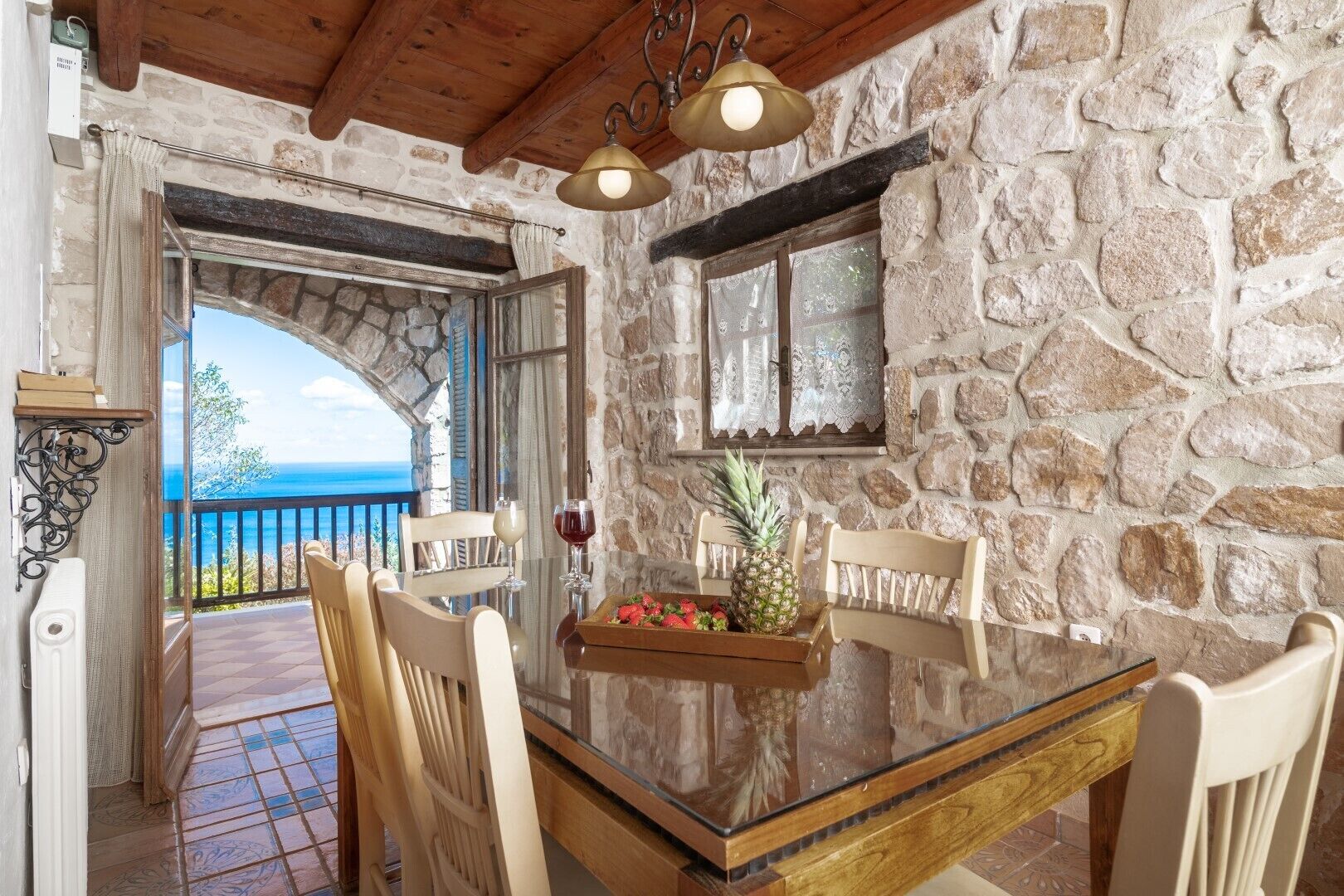 Una habitación con paredes de piedra, una mesa de vidrio, sillas de madera y vista al mar.