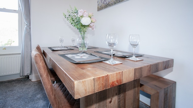 Una mesa de madera con un florero, dos copas de vino y un banco.