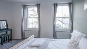 Un dormitorio con dos ventanas, una cama con ropa blanca, una mesita de noche con espejo y un radiador.