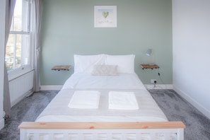 A neatly made bed with white linens and two towels, a bedside lamp, a small shelf, and a framed picture on the wall.