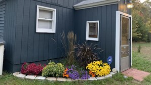 Una piccola casa di colore scuro con due finestre e una porta, circondata da una varietà di fiori colorati.