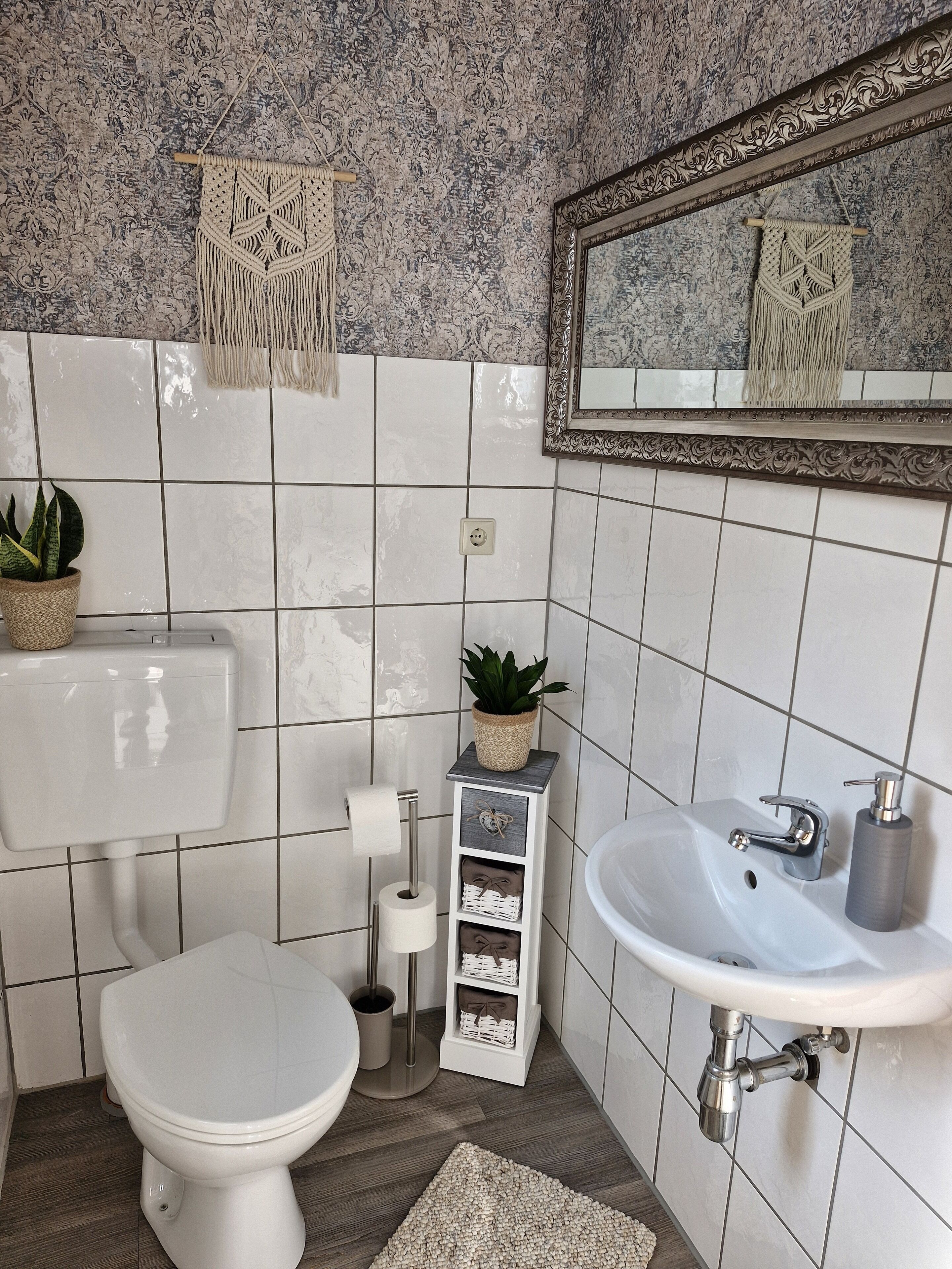 A bathroom with a toilet, sink, and mirror. There are decorative macramé hangings, a potted plant, and a small shelf with storage.