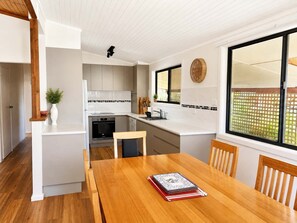 A modern kitchen with white cabinetry, a wooden dining table, and a large window with a view of greenery. - Lockhart Leafy Cottage (Mollymook)