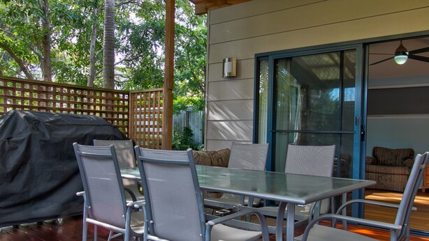 Un patio al aire libre con una mesa y sillas de vidrio, una parrilla cubierta y una puerta corrediza de vidrio que da a un espacio interior.