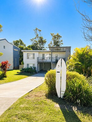 A modern two-story house with a visible garage door, a well-maintained lawn, and a surfboard leaning against the house.
