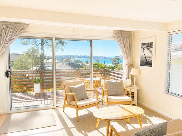 A room with a large sliding glass door leading to an outdoor deck with a view of the ocean and hills. Inside, there are two wooden chairs with cushions, a small round table, and a lamp on a side table.