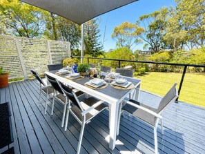 An outdoor dining area with a table set for four, surrounded by chairs, on a wooden deck with a view of a garden and trees. - Sea Breeze On Carroll (Mollymook)