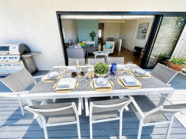 An outdoor dining area with a table set for six, surrounded by white chairs, on a wooden deck. - Sea Breeze On Carroll (Mollymook)