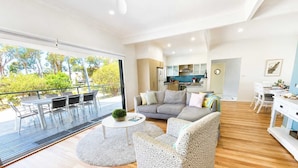 A modern living room with a sofa, armchair, and coffee table, a balcony with outdoor furniture, and a kitchen in the background. - Sea Breeze On Carroll (Mollymook)