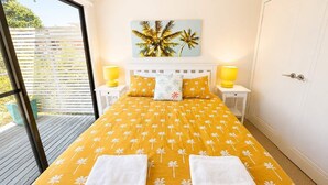 A bedroom with a bed, two bedside tables, a painting, and a view through a glass door. - Sea Breeze On Carroll (Mollymook)