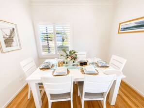 A dining room with a white table set for four, white chairs, and a window with blinds. - Sea Breeze On Carroll (Mollymook)
