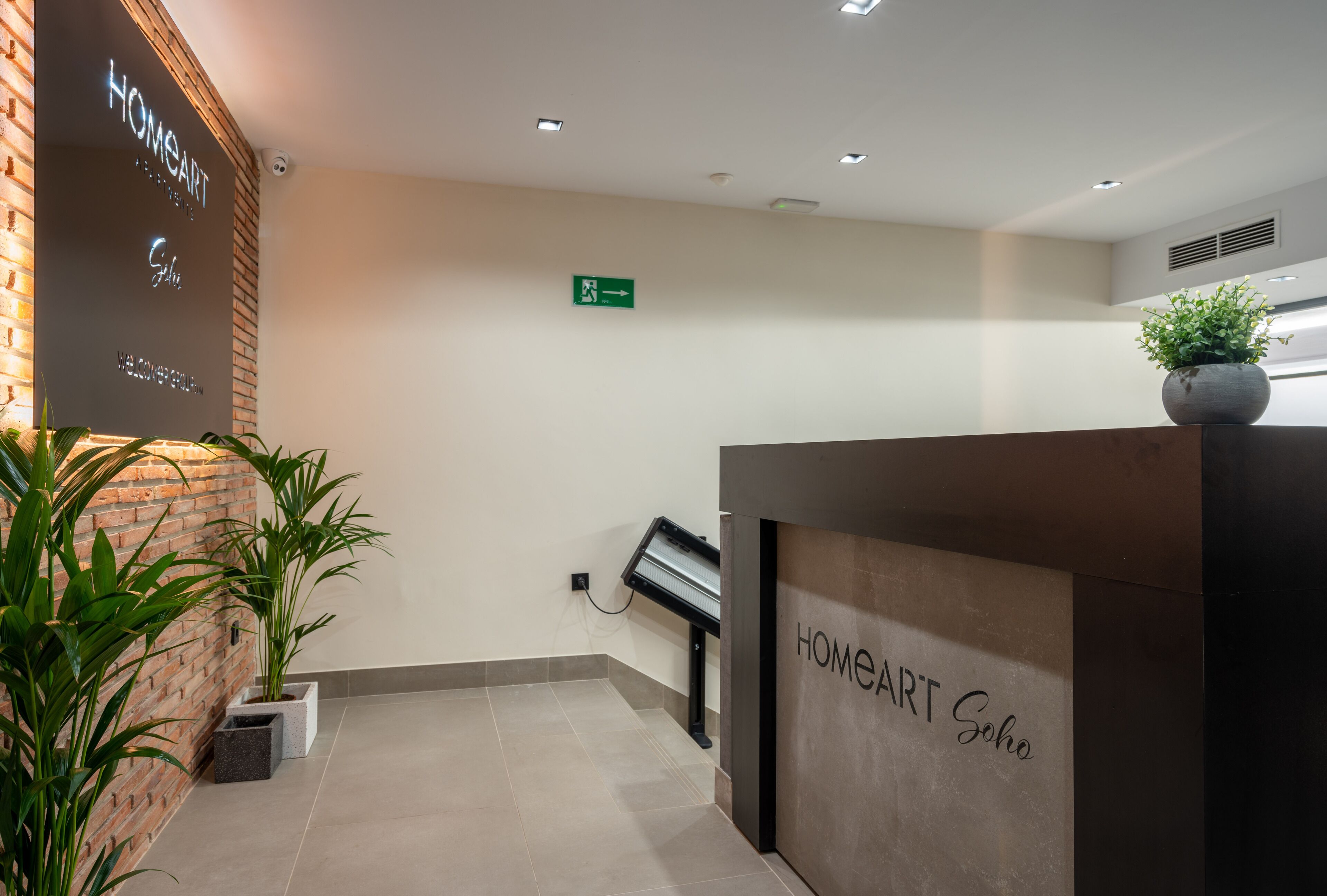 A reception area with a brick wall, a reception desk labeled "HOMEART Soho," and a green emergency exit sign.