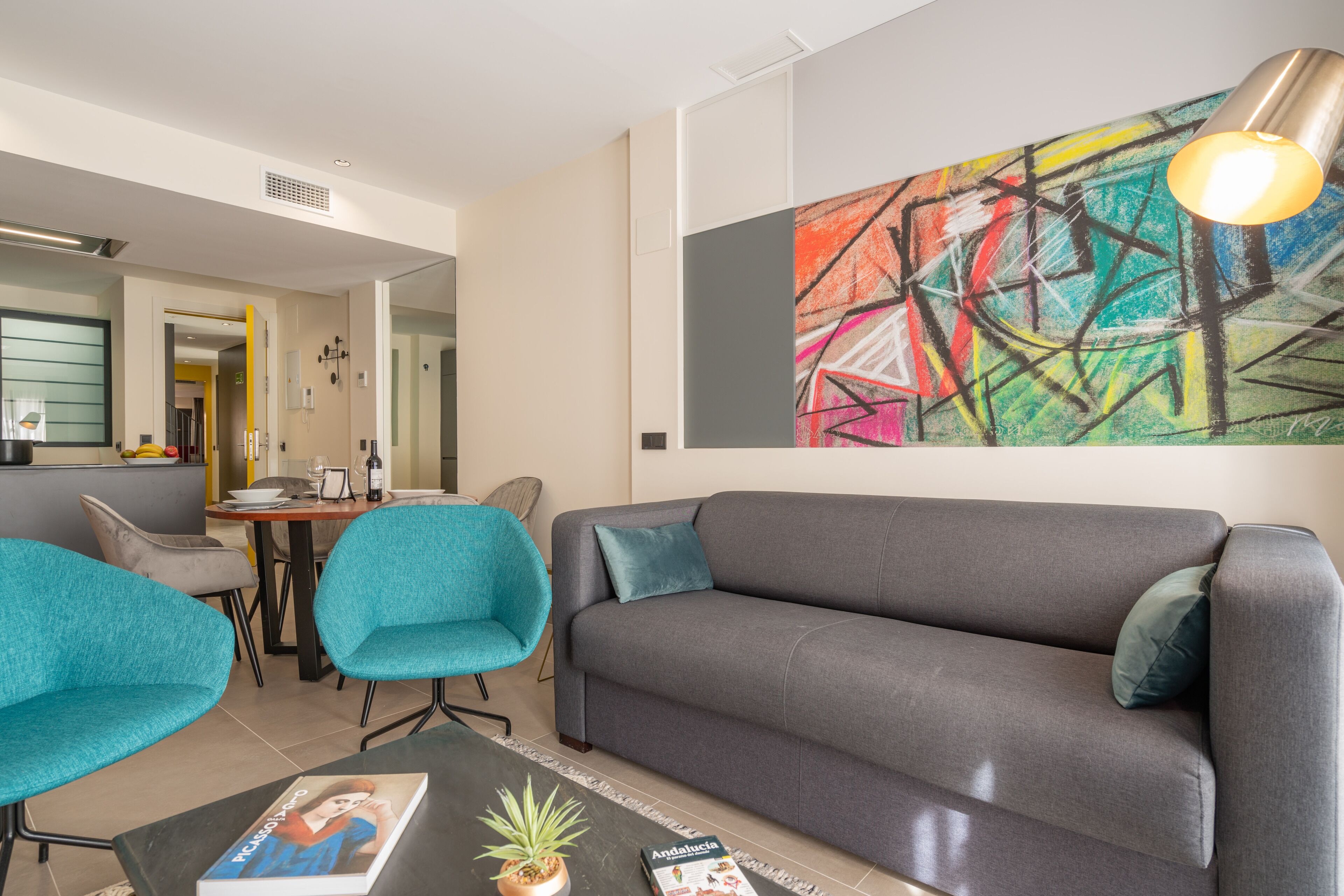 Ein modernes Wohnzimmer mit einem grauen Sofa, türkisfarbenen Sesseln und einem farbenfrohen abstrakten Gemälde an der Wand.