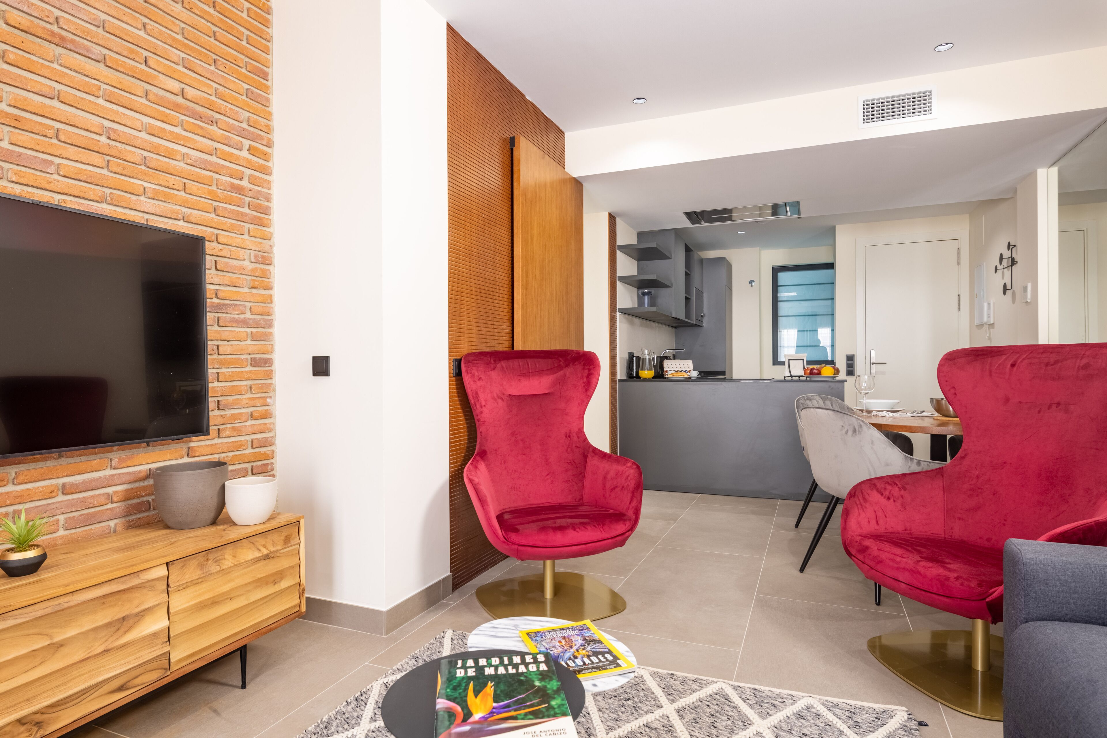 A modern living room with a brick wall, a flat-screen TV, a wooden console, red and grey chairs, and a dining area with a grey table and chairs.