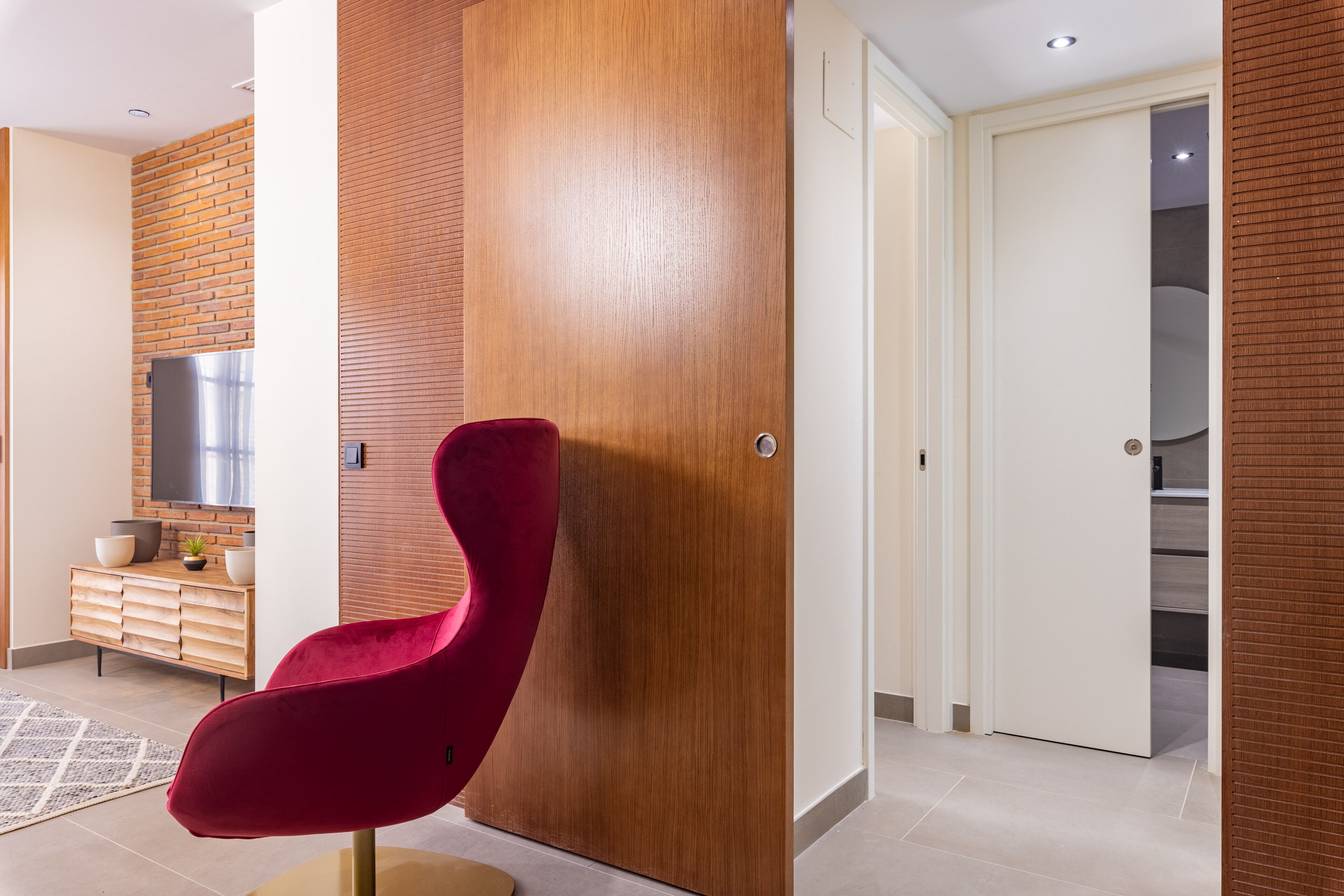 A modern interior with a red chair, wooden panel walls, a kitchen area with a refrigerator, and a white door leading to another room.