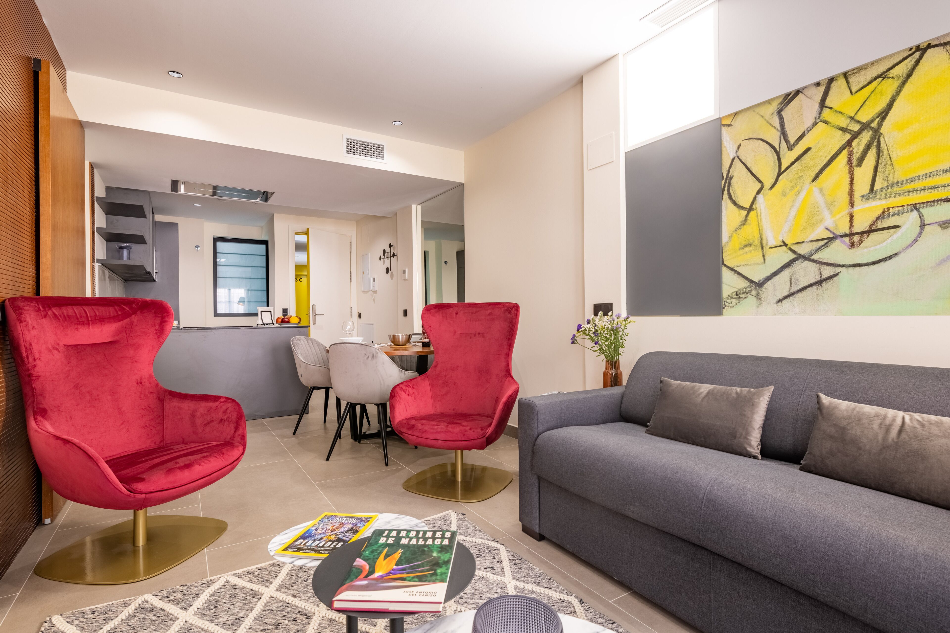 Ein modernes Wohnzimmer mit einem grauen Sofa, roten Sesseln und einem Essbereich mit Tisch und Stühlen.