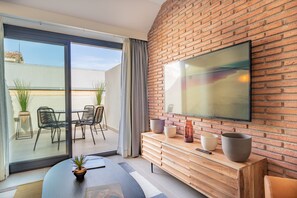Een moderne woonkamer met een grote televisie, een houten dressoir en een glazen schuifdeur die leidt naar een buitenruimte met stoelen en planten.