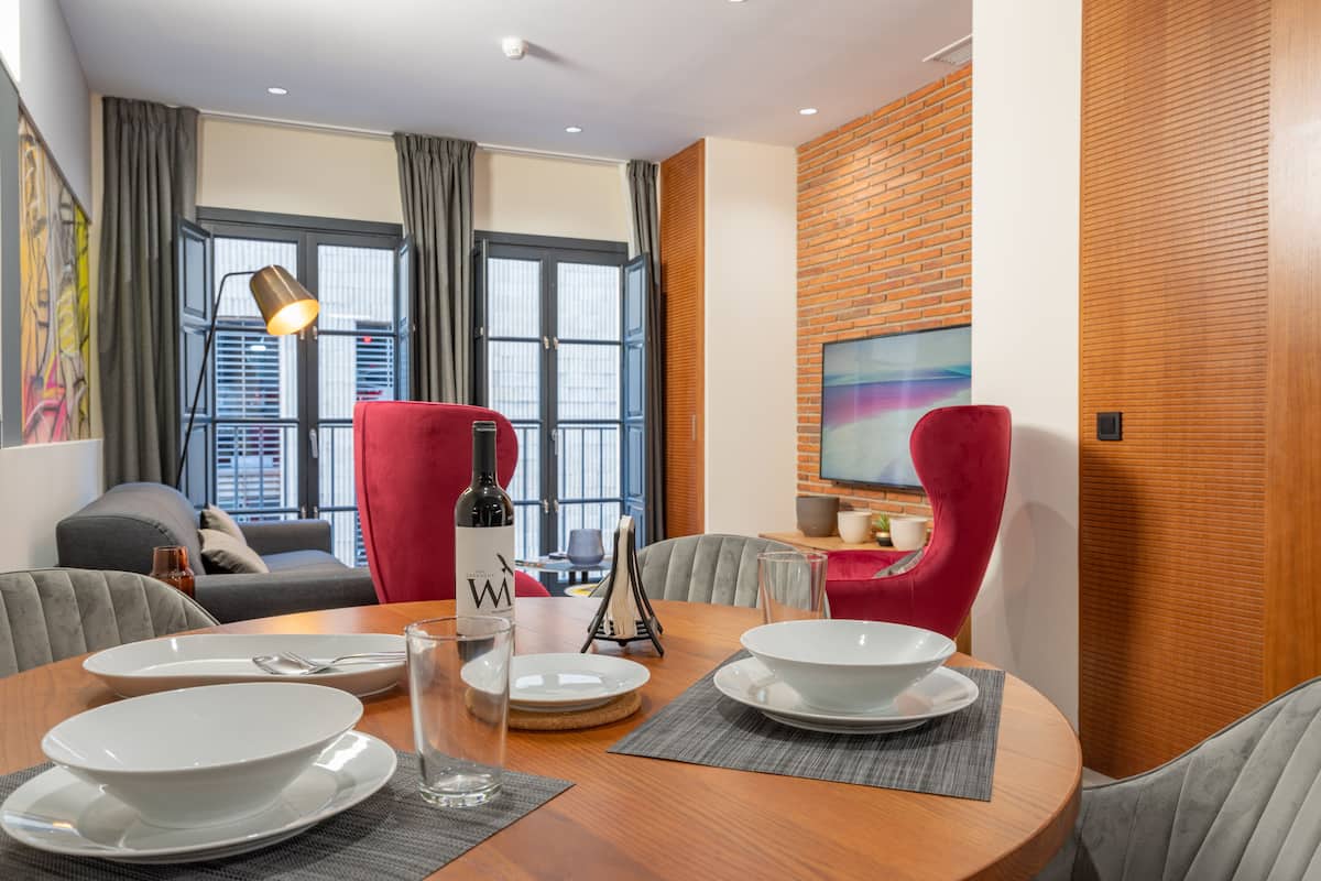 A modern dining area with a round wooden table set for two, two red chairs, a wine bottle, and a painting on the wall.