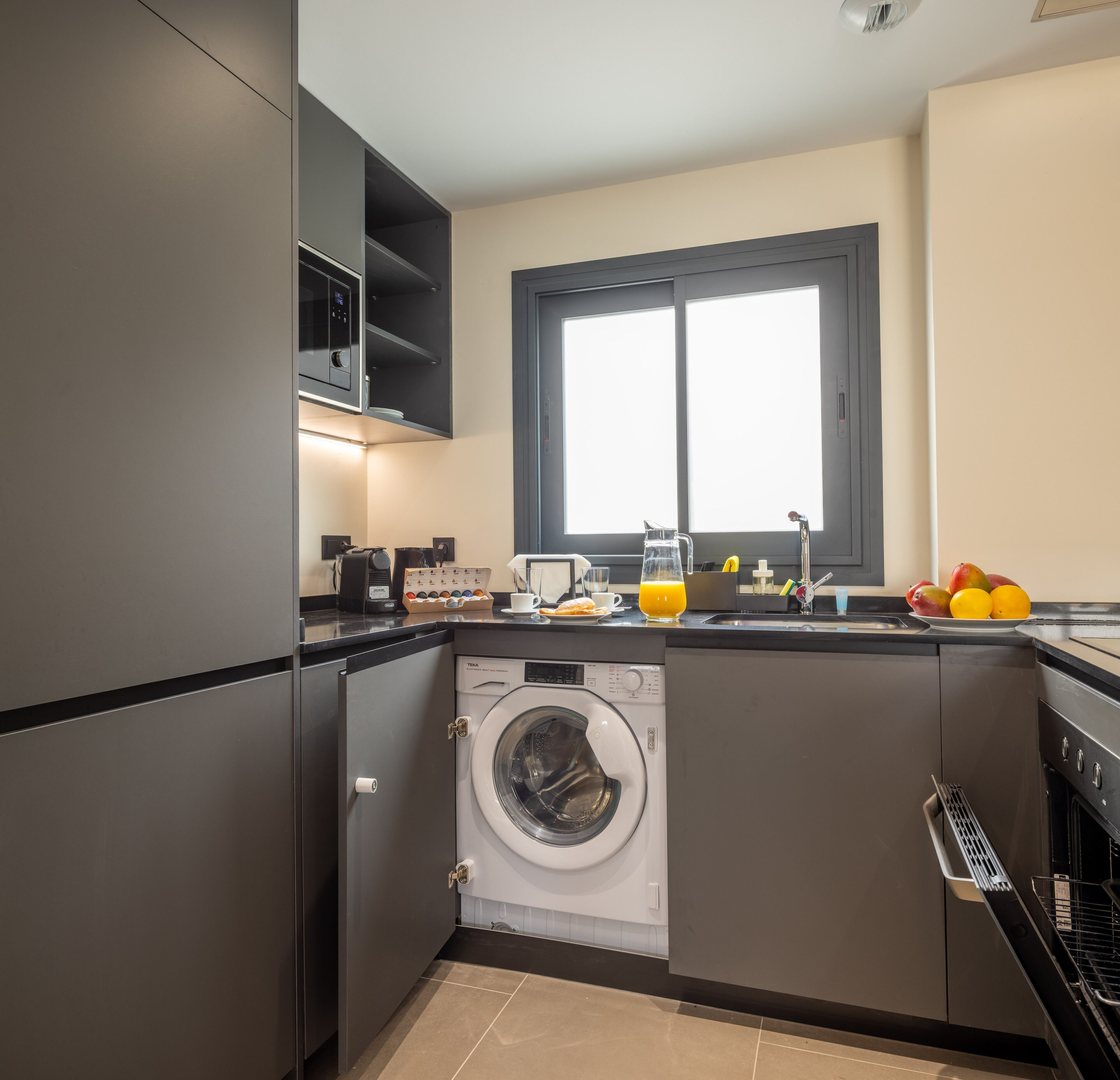 A compact kitchen with a washing machine, a coffee maker, and a fruit bowl on the counter.