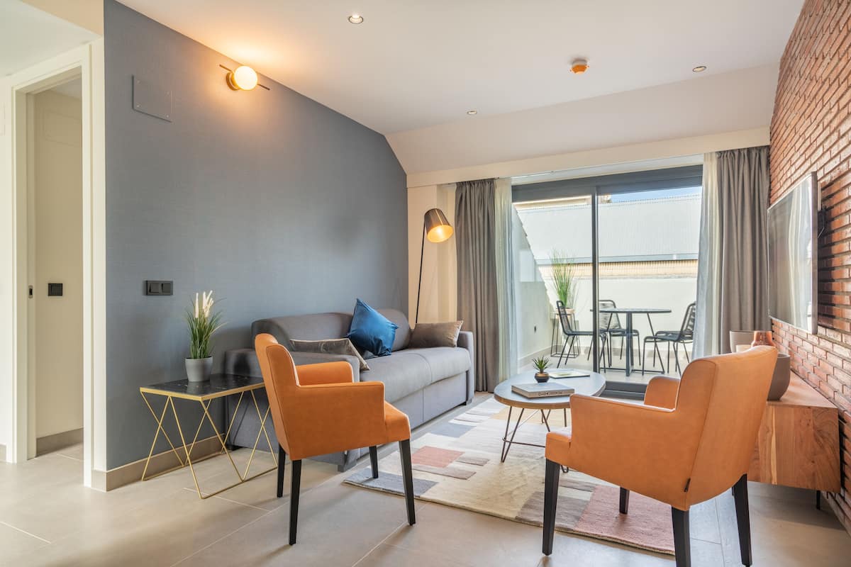 A modern living room with a grey accent wall, a grey sofa, orange chairs, a small table, and a view of the outdoors through large windows.