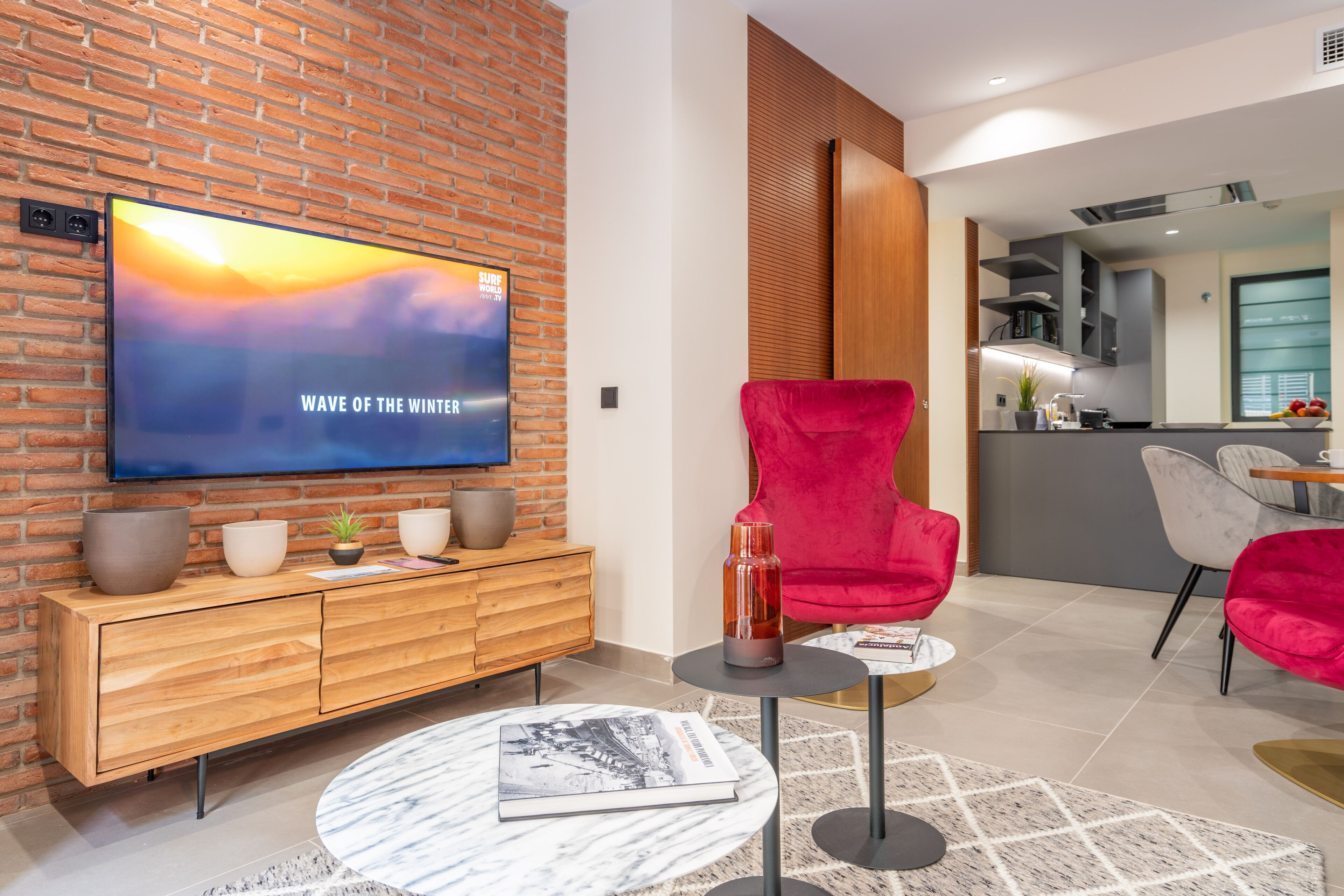 A modern living room with a brick wall, a wooden cabinet, a red chair, a round coffee table, and a flat-screen TV displaying "WAVE OF THE WINTER".