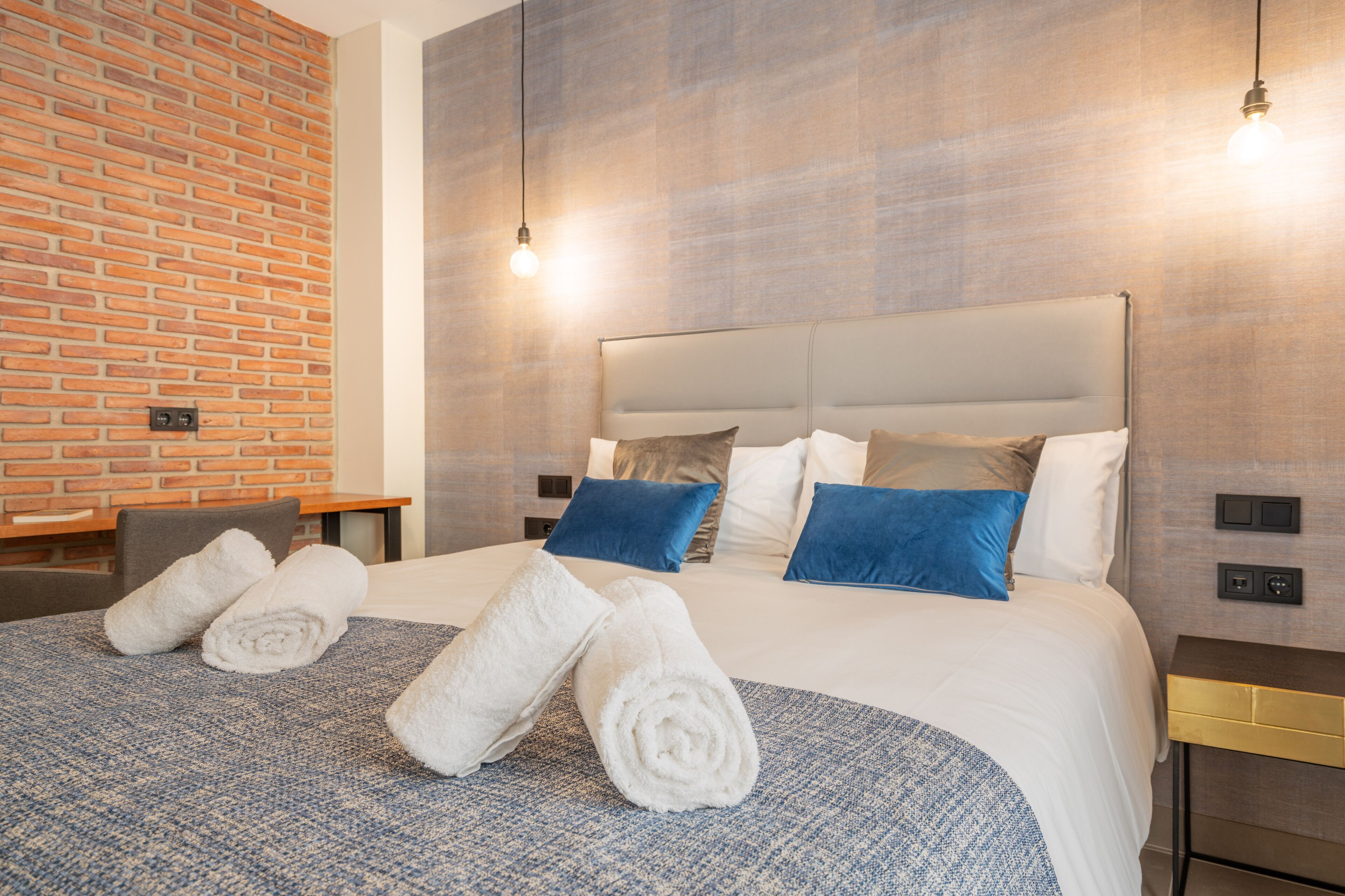 Habitación de hotel con pared de ladrillo, una cama grande con ropa de cama azul y blanca, y dos toallas enrolladas sobre la cama.