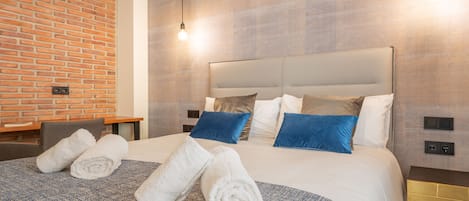Habitación de hotel con pared de ladrillo, una cama grande con ropa de cama azul y blanca, y dos toallas enrolladas sobre la cama.