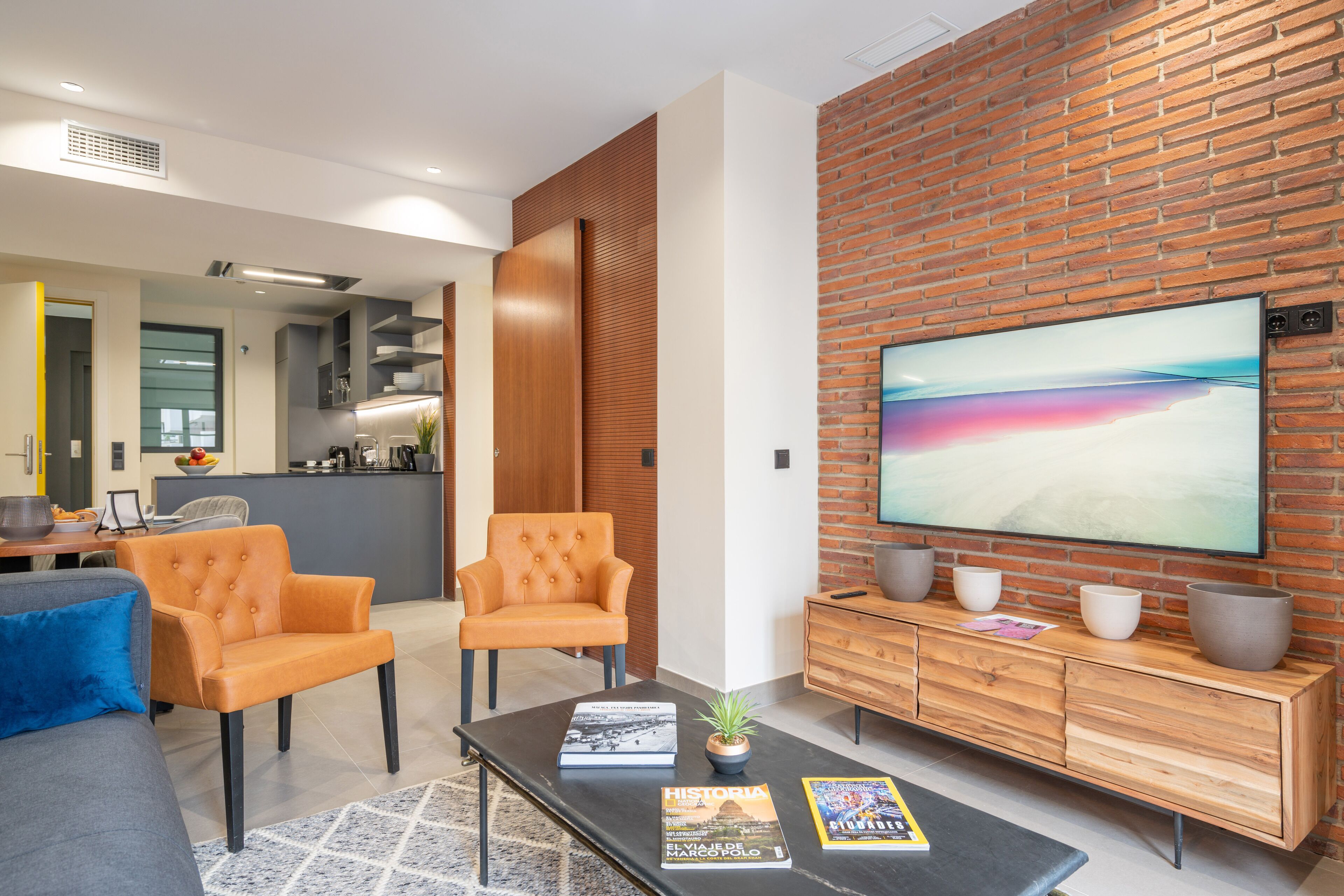 Ein modernes Wohnzimmer mit Ziegelwand, großem Flachbildfernseher, zwei orangefarbenen Sesseln, einem Couchtisch mit Büchern und einem Holzkabinett.
