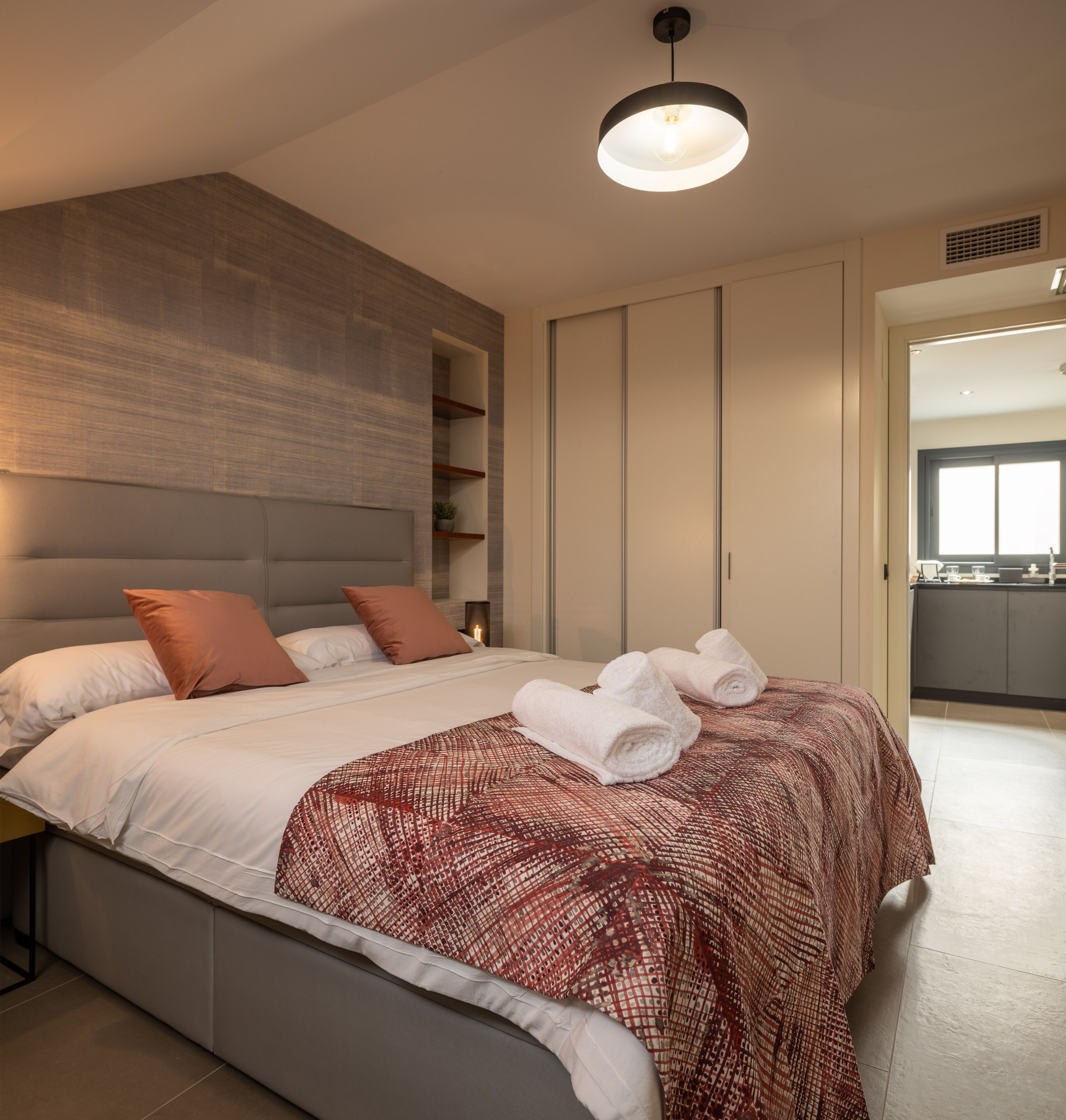 Een moderne slaapkamer met een groot bed, hoofdeinde, nachtkastje, wandplank en plafondlamp.
