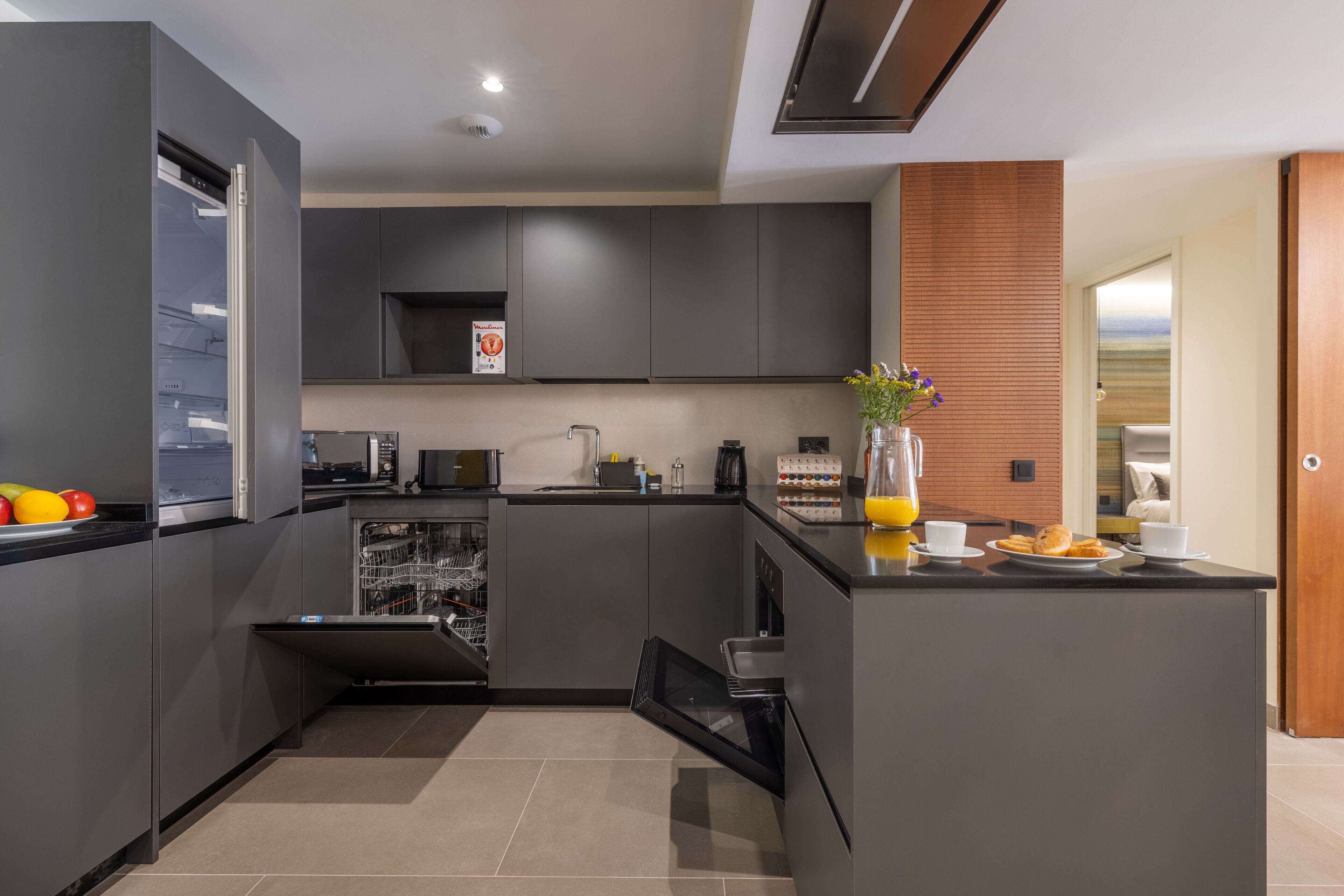 A modern kitchen with grey cabinets, a built-in dishwasher, and a breakfast bar with a vase of flowers and a plate of croissants.