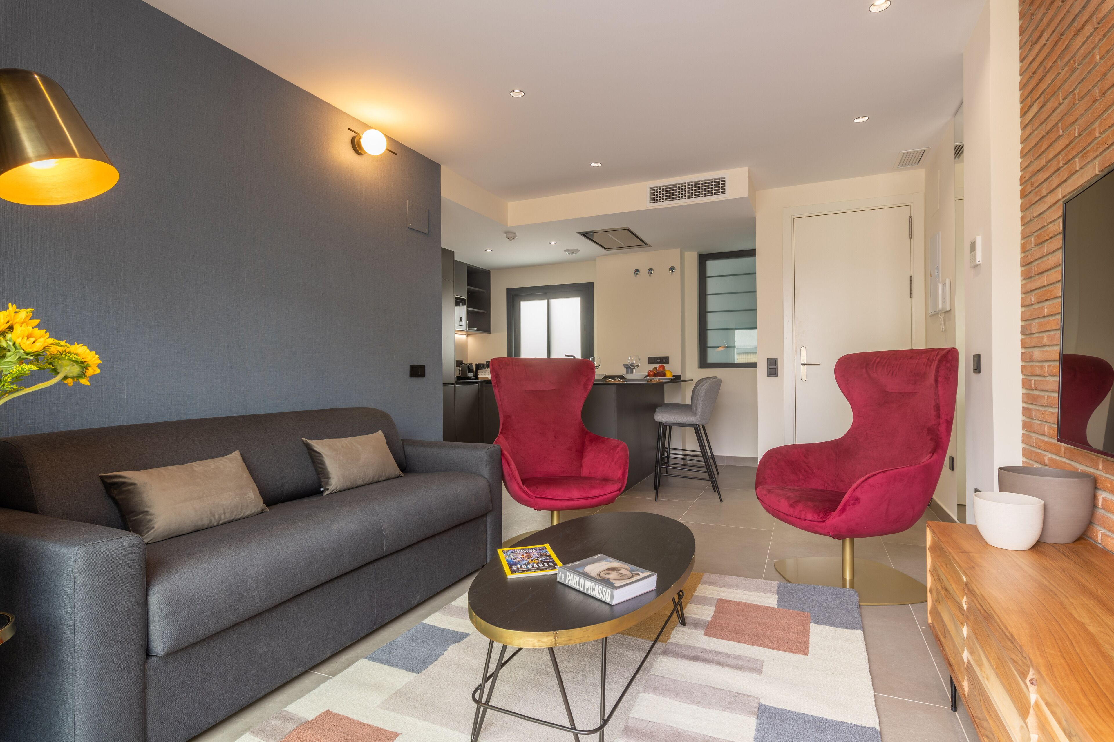 Una sala de estar moderna con un sofá gris, dos sillas rojas, una mesa de centro y una zona de cocina al fondo.