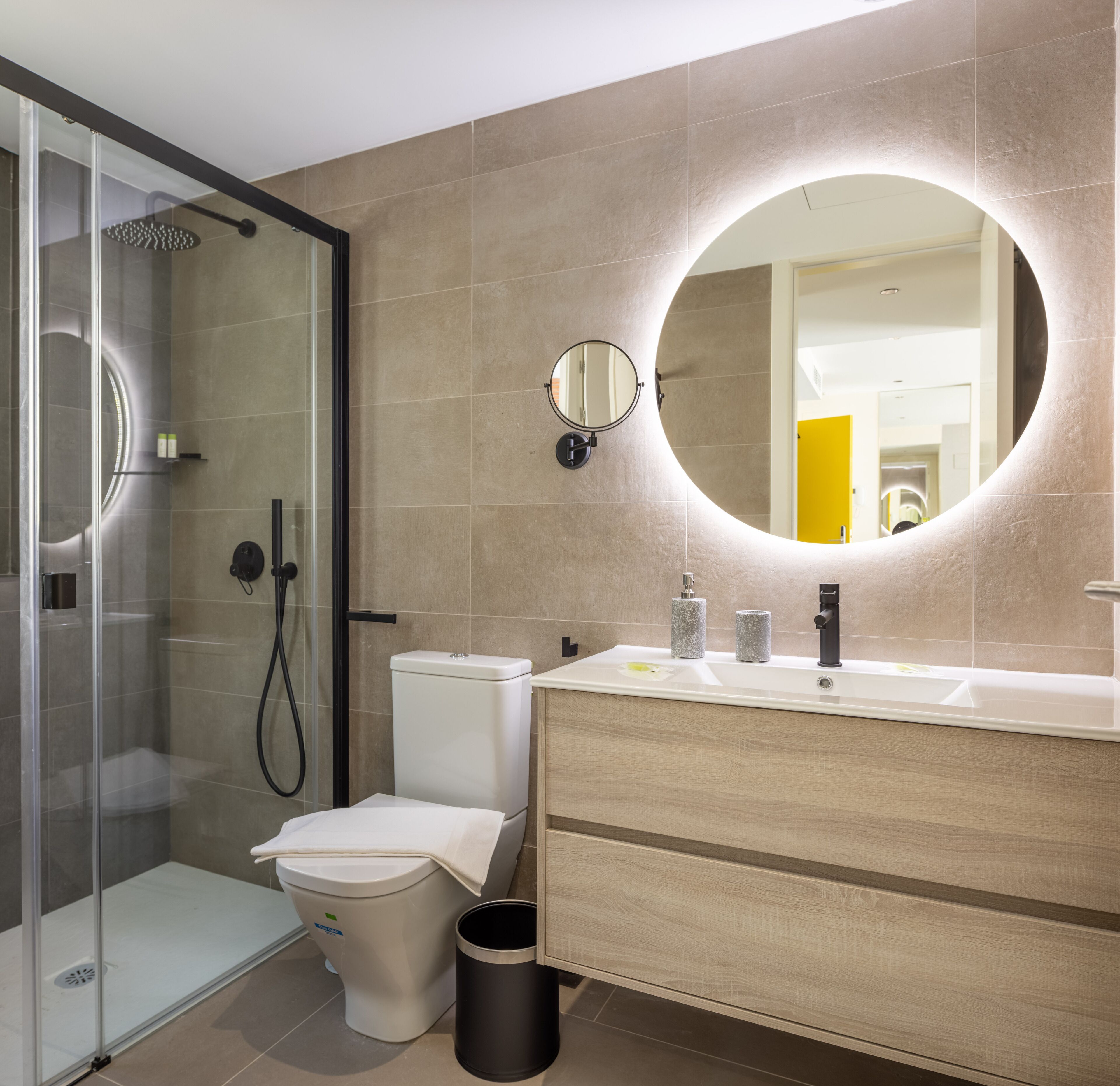 Un baño moderno con ducha de vidrio, inodoro, lavamanos con espejo y un cesto de basura.