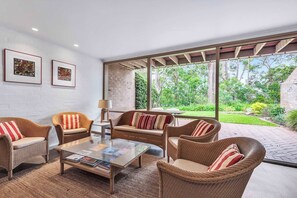 Ein modernes Wohnzimmer mit Rattanmöbeln, einem Glastisch und großen Fenstern mit Blick auf einen Garten.