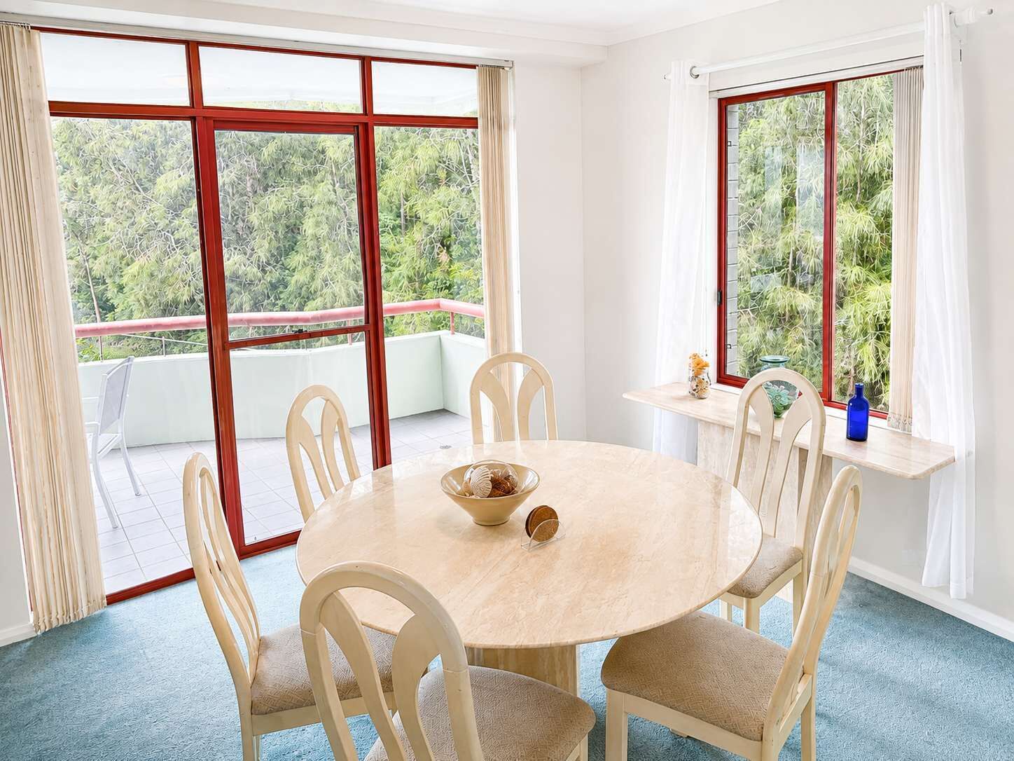 Mesa de jantar redonda com seis cadeiras, janelas amplas com molduras vermelhas e vista para o verde lá fora.