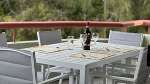 A white table set outdoors with a bottle of wine, glasses, and woven placemats.