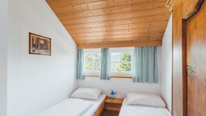 A small, cozy room with two single beds, a wooden ceiling, a window with curtains, and a small table with a lamp.