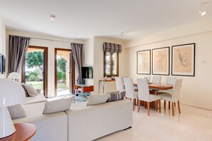 Uma sala de estar moderna com sofá, mesa de jantar e cadeiras. Há grandes janelas com cortinas, uma televisão de tela plana e quadros na parede.