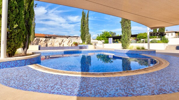 A swimming pool with blue mosaic tiles, surrounded by a covered area with trees and a view of a building in the background. - Apartment A02 - Roudias (Kouklia)