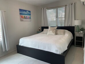 A modern bedroom with a large bed, a nightstand with a lamp, and a painting of beach chairs. - Malecon Beach House with beautiful backyard. Steps from the Caribbean ocean. (PATILLAS)