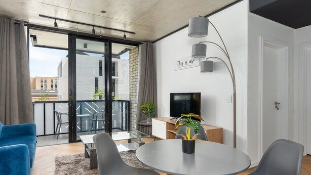 A modern living room with a round table, grey chairs, a blue sofa, a TV, and a balcony with a view of buildings. - 1 Bedroom on Century City Urban Square (Cape Town)