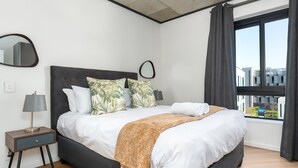 Una habitación de hotel moderna con una cama grande, una mesita de noche con lámpara, un espejo y una ventana con cortinas.