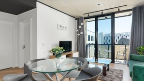 Una sala de estar moderna con una mesa de comedor de vidrio, sillas grises, un sofá verde, un televisor de pantalla plana y un balcón con vista a la ciudad.