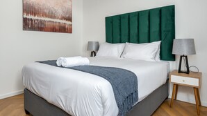 A neatly made bed with white linens and a green headboard, a grey throw blanket, and a small bedside table with a lamp. - Bridgewater One  | Netflix | Fibre (Cape Town)