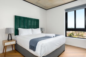 A modern bedroom with a large bed, a green headboard, a nightstand with a lamp, and a window with a city view.