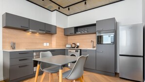 A modern kitchen with grey cabinets, a stainless steel refrigerator, a built-in washing machine, and a dining area with grey chairs and a wooden table. - Bridgewater One  | Netflix | Fibre (Cape Town)