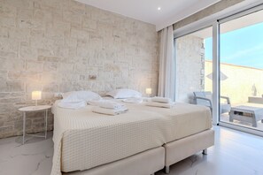 A bedroom with a stone wall, a large bed with white linens, a small bedside table with a lamp, and a view through a glass door to an outdoor seating area.