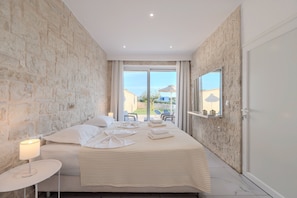 Un dormitorio con pared de piedra, una cama con ropa blanca, una mesita de noche con lámpara y vistas al exterior a través de un ventanal.