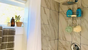 A bathroom with a shower, marble tiles, a towel rack with towels, and toiletries on a shelf. - The Indigo House at Canyon Lake (Canyon Lake)