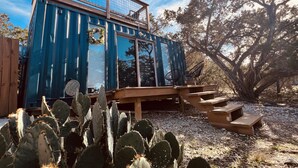 A modern, blue, metal-framed structure with a wooden deck and benches, surrounded by cacti and trees. - The Indigo House at Canyon Lake (Canyon Lake)