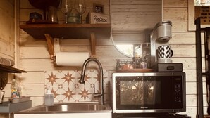 A compact kitchen with a sink, a microwave, and a refrigerator. There is a shelf above the sink with various items, and a mirror on the wall. - The Indigo House at Canyon Lake (Canyon Lake)